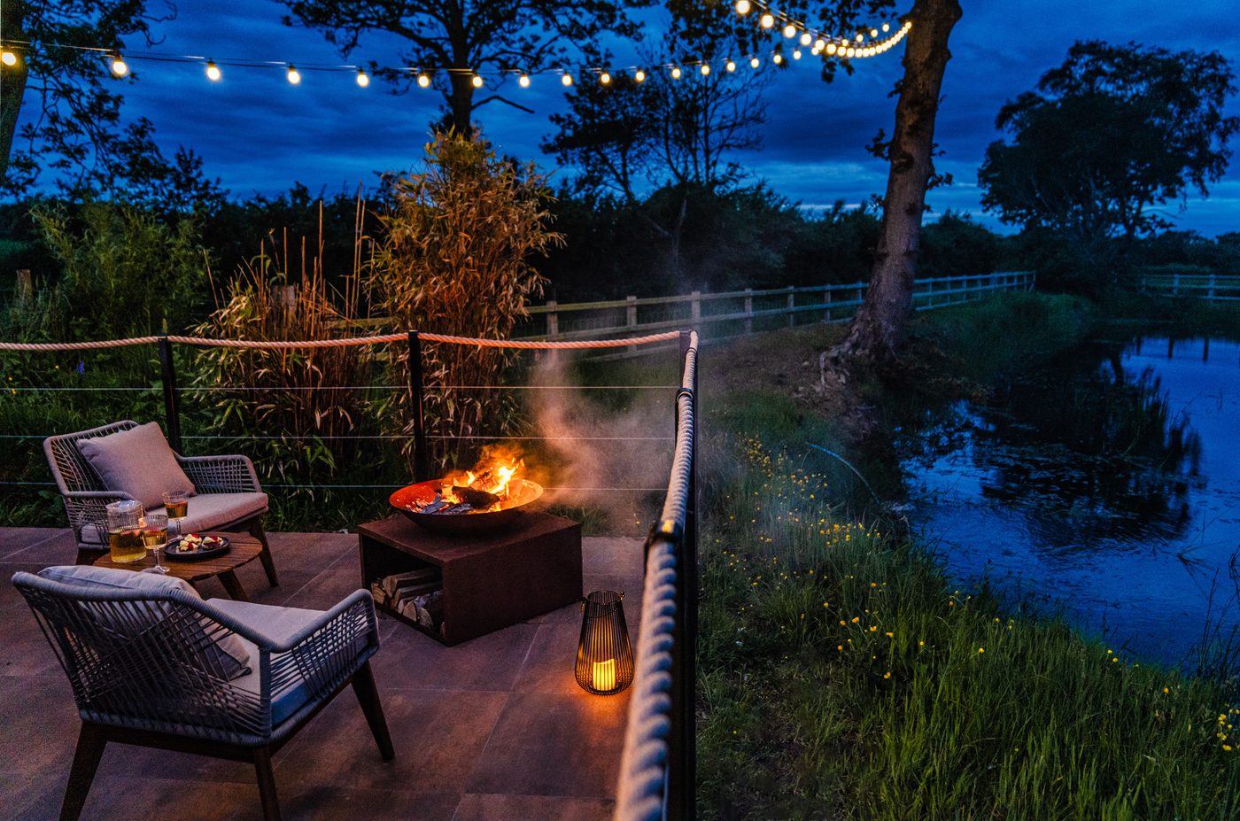 Fire bowl blazing on the Rusty Boathouse terrace at dusk — fairy lights and the lake