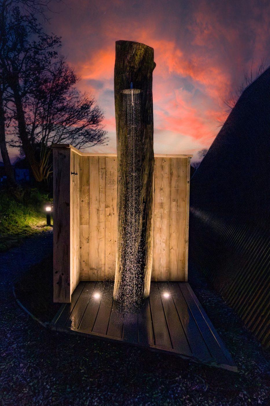 Outdoor tree-trunk shower at the Tri-Pod lit at night under a dramatic Welsh sky