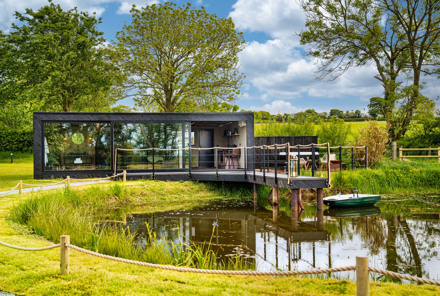 The Rusty Boathouse exterior in daylight — dark-clad cabin over the lake with rowing boat